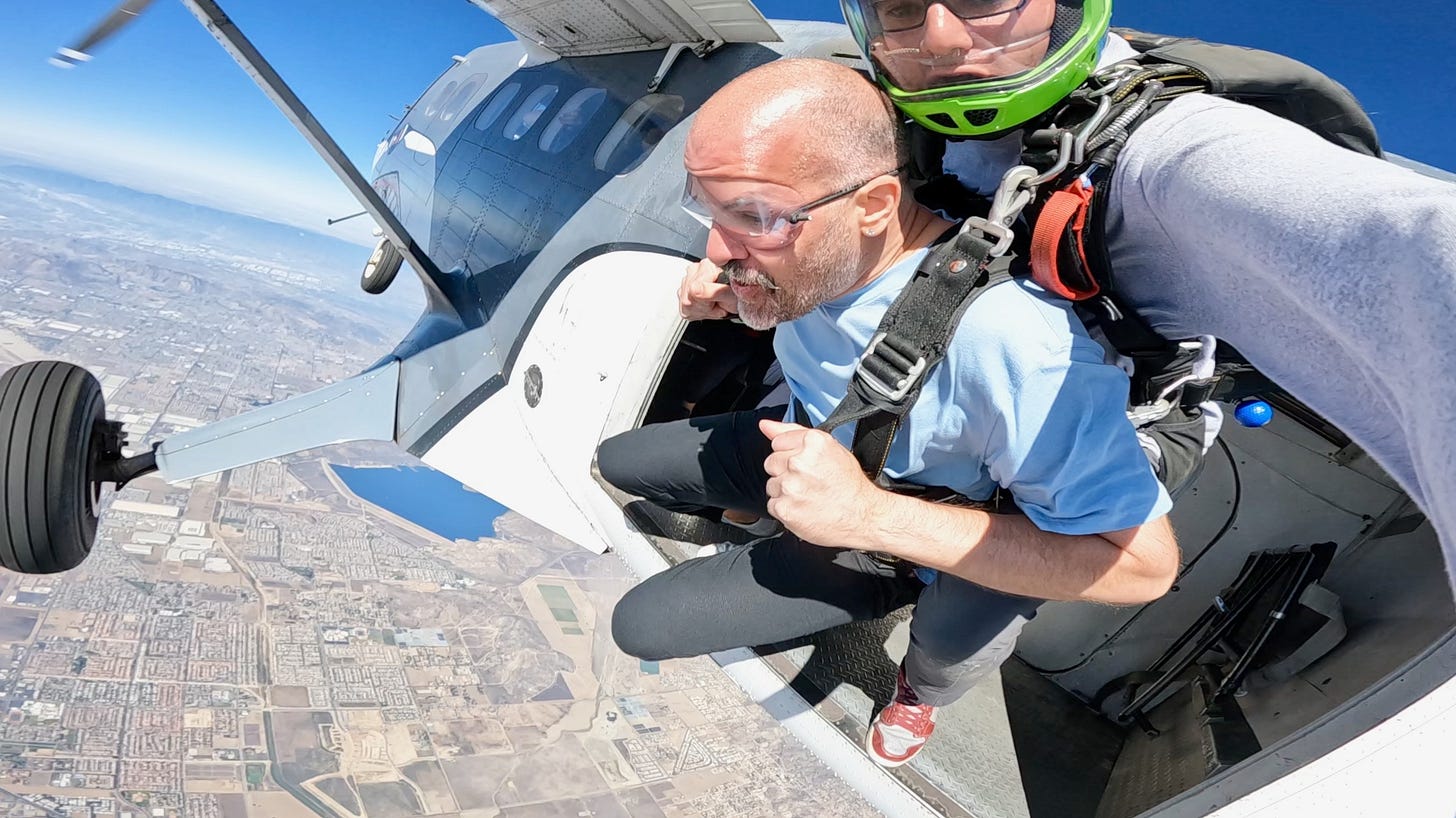 Mid-tandem skydive at 18,000 feet: the author in a blue shirt hanging out of the plane’s door with his instructor, Earth below.