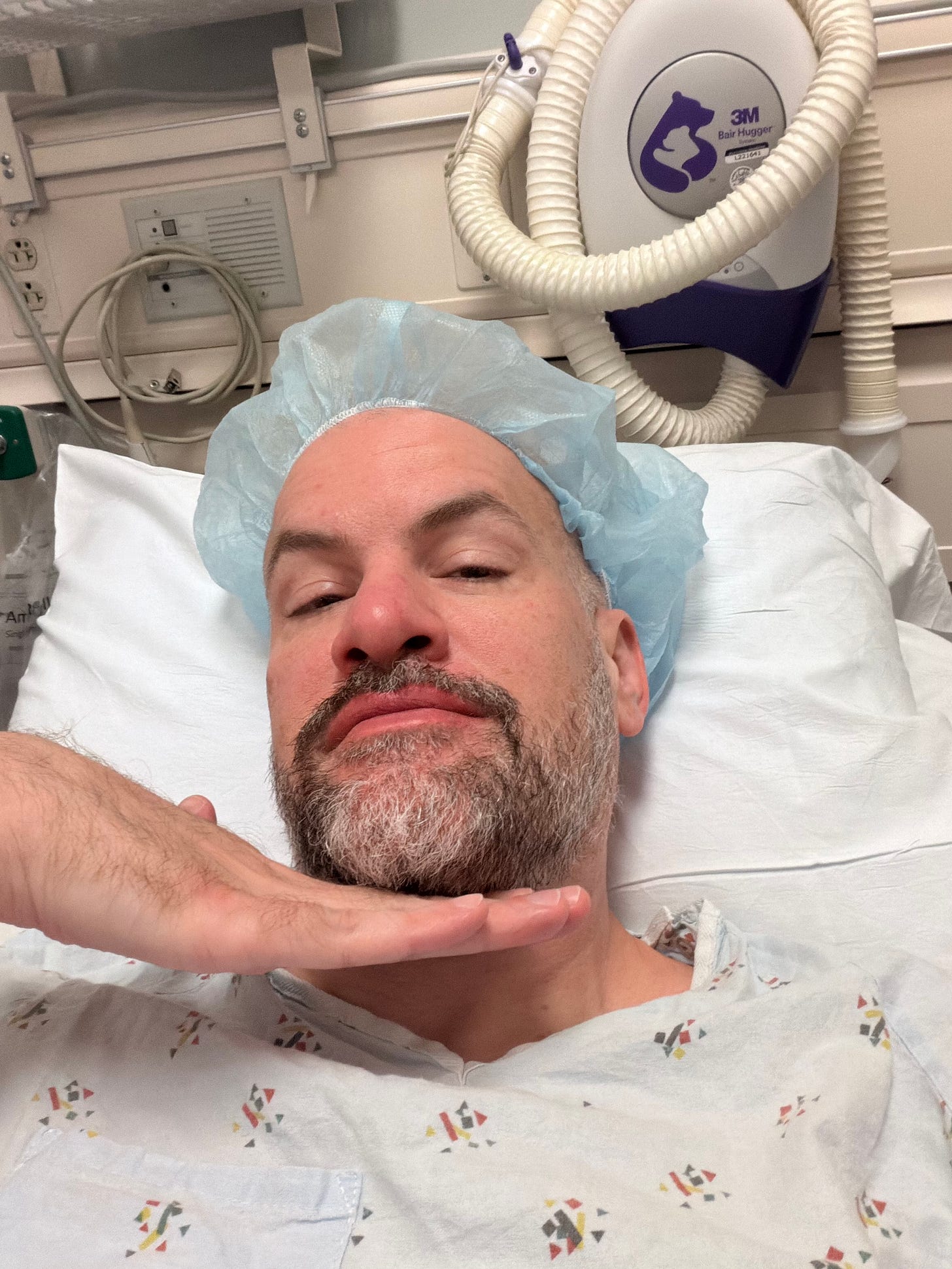 Selfie from the hospital bed on surgery morning: blue cap, gown, game face on.